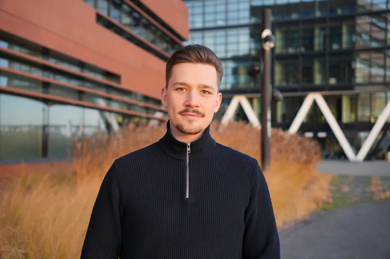 Tom Falz im dunklen Pullover vor einem Bürogebäude, im Vordergrund herbstliche Gräser